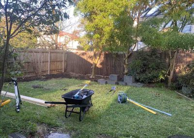 Before: grassy backyard with a wheelbarrow, tools and blocks, wider view